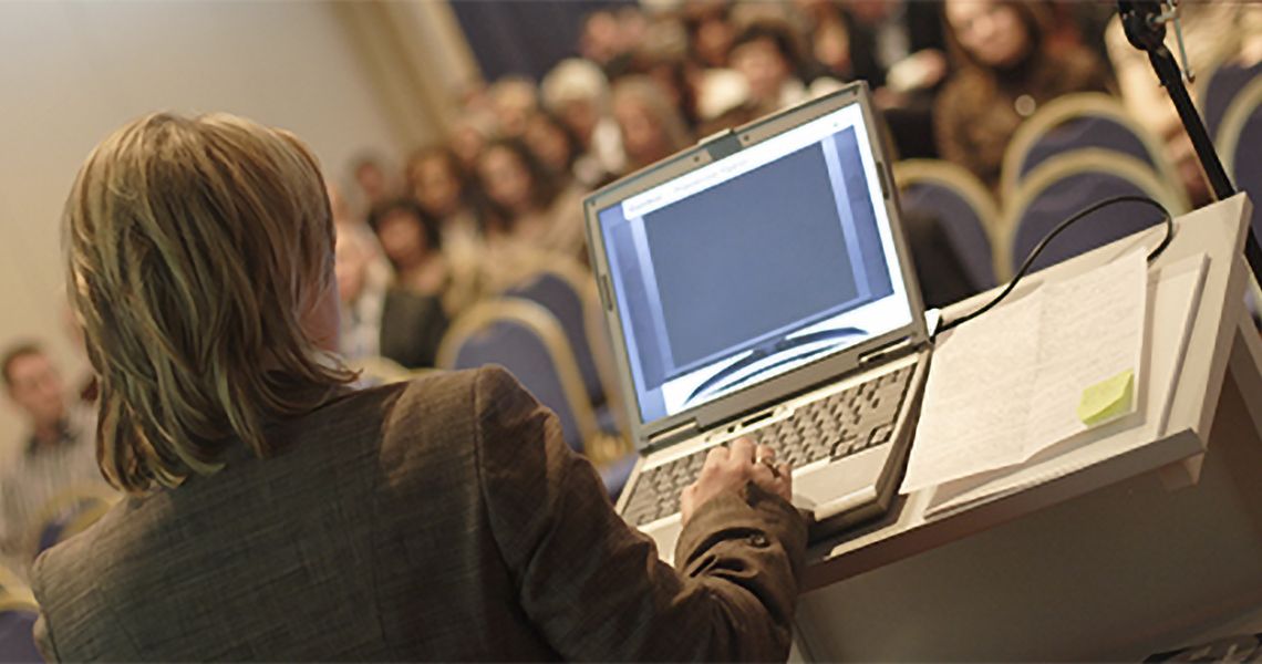 Person standing at a podium looking at a laptop, with an audience looking on